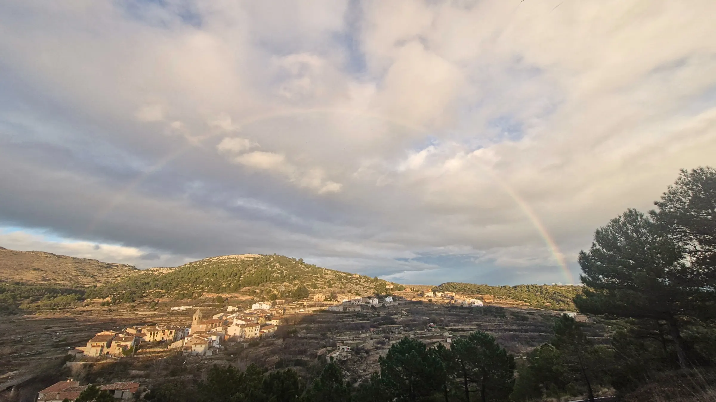 Fuentes de Rubielos sobre la ladera, rodeado de colinas boscosas, bajo un cielo nuboso con un arcoíris