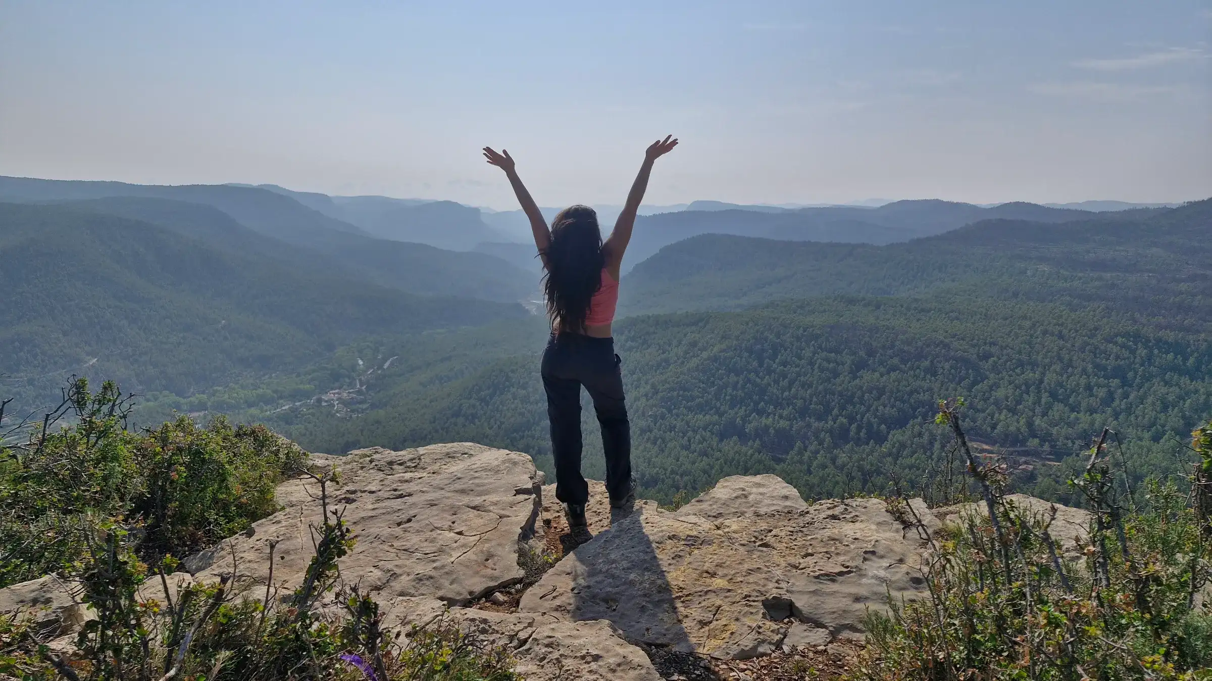 Montañera de espaldas con los brazos en alto sobre un valle boscoso